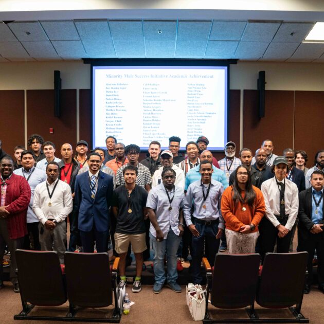 A large group of approximately 35 minority male students standing in three rows in an auditorium with a screen behind them.