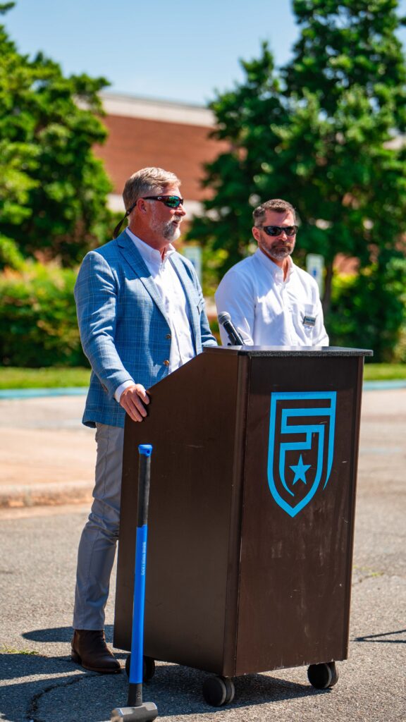 Two men, both wearing sunglasses, stand outside. One is speaking at a podium. 