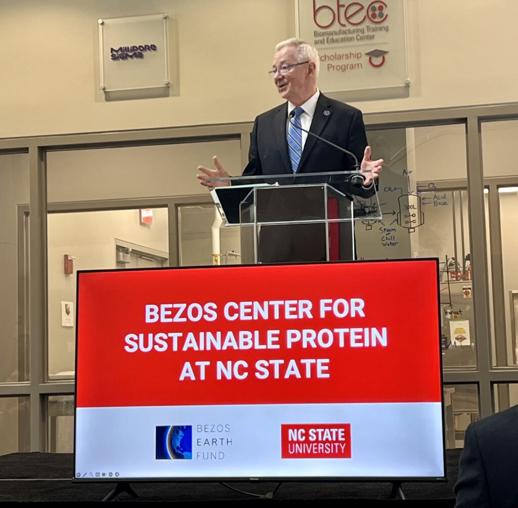 A man with grey hair and glasses in a dark suit with a blue striped tie stands in front of a podium with a screen in front. On the screen are the words "Bezos Center for Sustainable Protein at NC State" with a logo for the Bezos Earth Fund and NC State University underneath. 