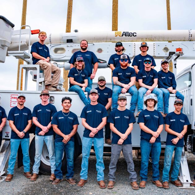 Large group of men wearing blue shirts, standing in front of a lineworker vehicle