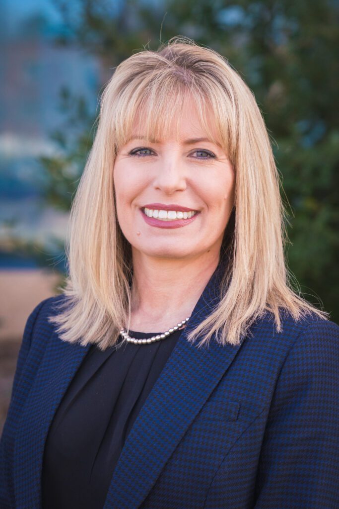 A photo of a woman with blonde hair and bangs. She is smiling and wears a navy blue blazer with a dark shirt underneath. 