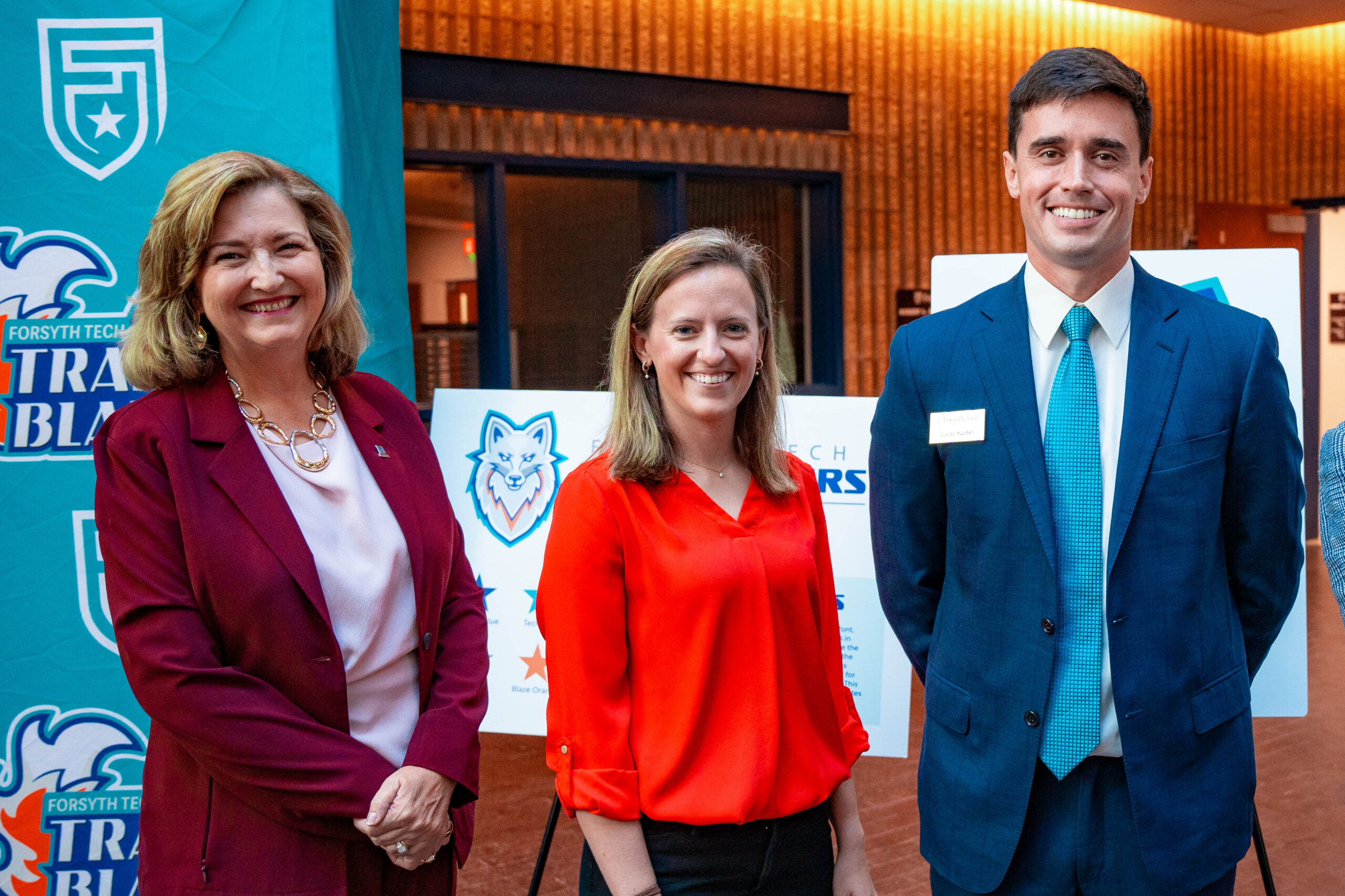 Three people stand smiling in a row. Far left is a woman wearing a maroon blazer with a white top underneath, with almost shoulder-length, dark blonde hair. The person in the middle is a woman wearing a red top with shoulder-length hair and the person on the right is a man with dark hair wearing a suit.