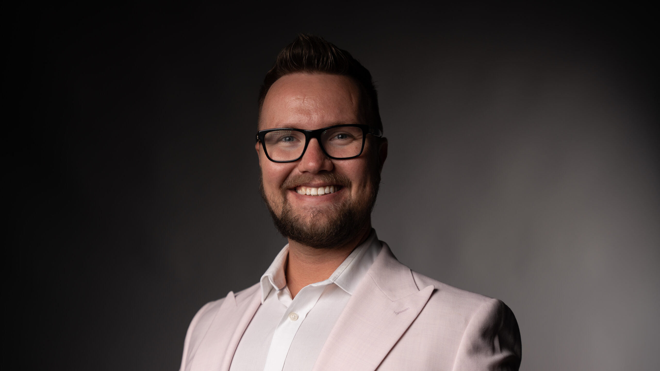 A close up photo of a smiling man, wearing glasses and a white collared shirt with light pink blazer. He is smiling and has facial hair. 