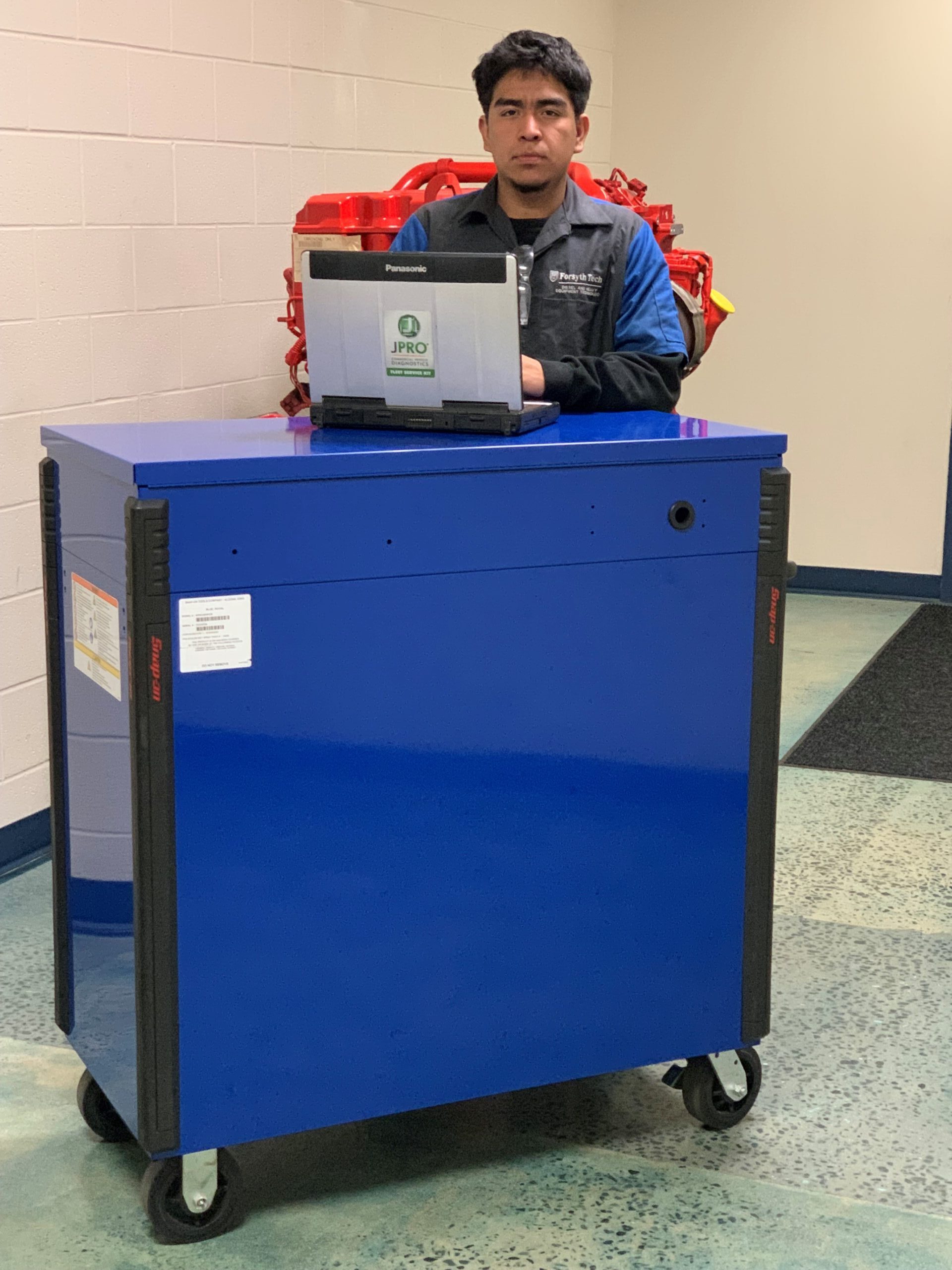 A man with dark hair stands behind an open laptop that is perched on a large, blue toolbox on wheels. The man is wearing a dark grey (almost black) and blue collared shirt with a dark, long-sleeved shirt underneath. 