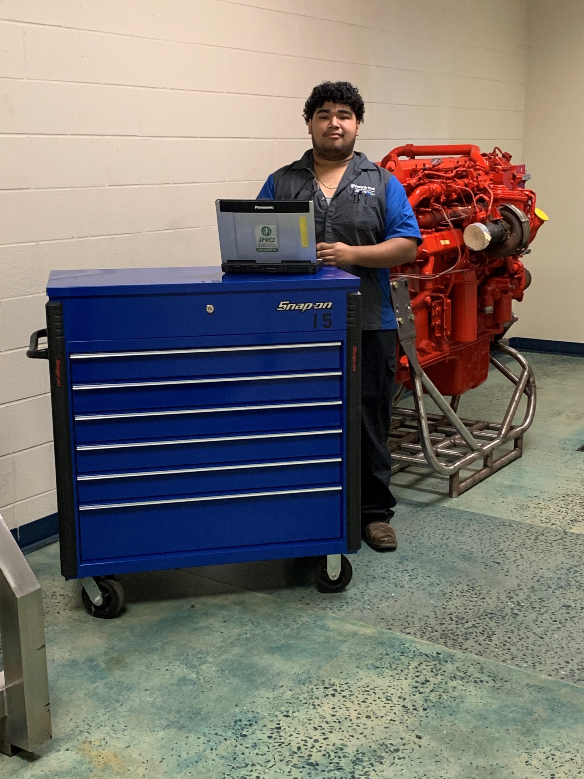 A male with brown skin and dark hair stands in front of a blue toolbox on wheels. He is wearing a collared shirt that is dark grey and blue and a large diesel engine is behind him. 