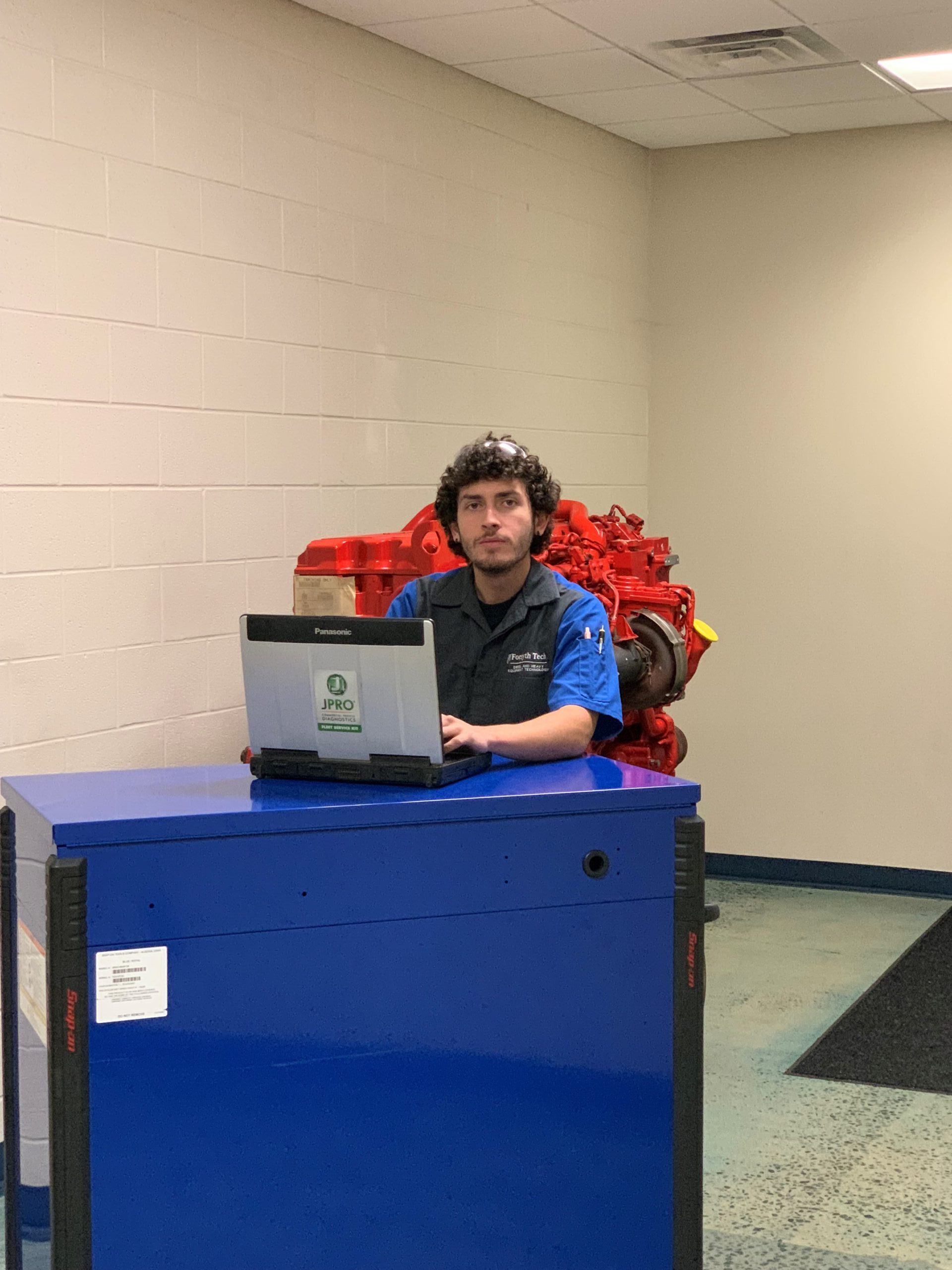 A male is posed behind a laptop. The laptop is on top of a blue toolbox; the man is wearing a blue and dark grey collared shirt and there is a large, red diesel engine behind him. 