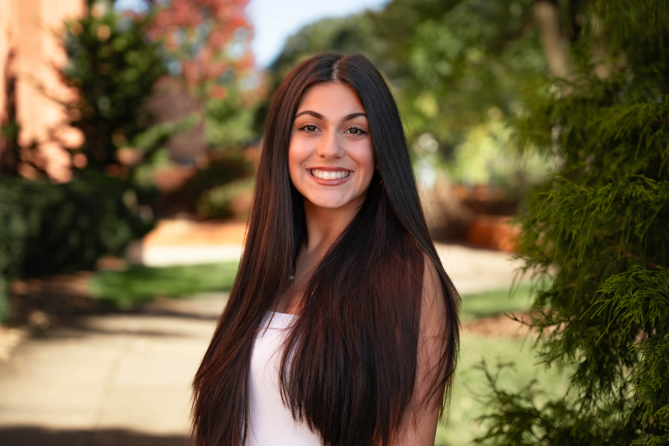 A young, dark-haired woman is smiling and facing the camera. She is outside on a sunny day with a sidewalk and dark plants in the background. 