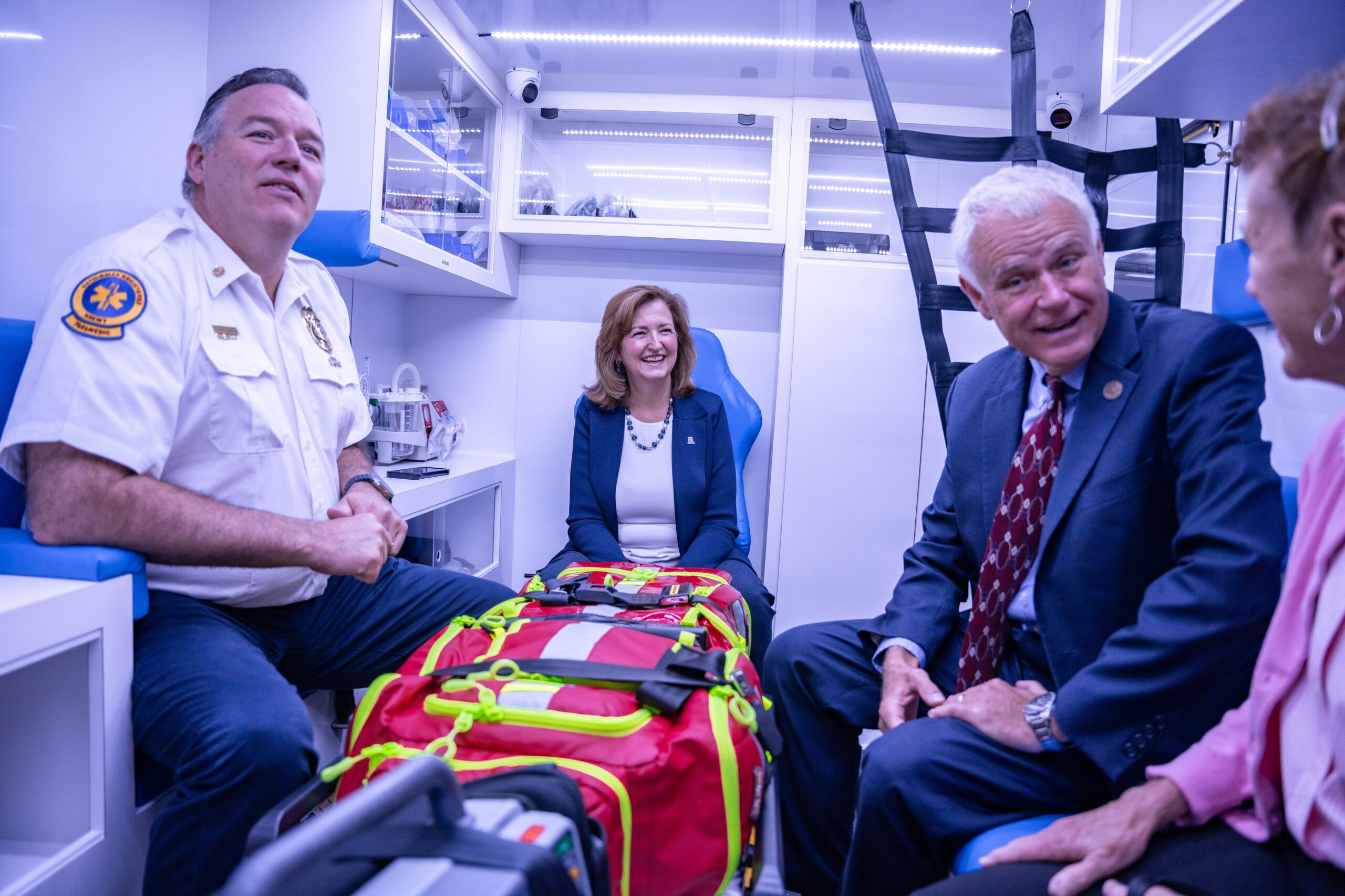 four people sit inside of an ambulance simulator; there is a gurney with a red piece of fabric over it with yellow reflective bands